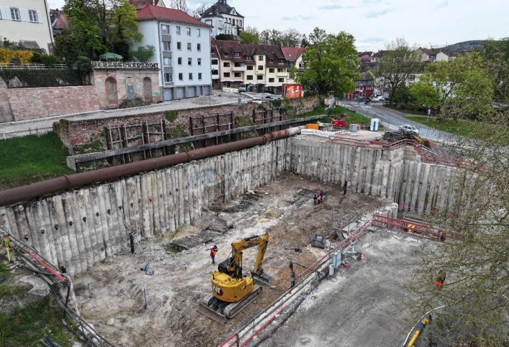 An Iron Age structure in Aschaffenburg has emerged from beneath the bank of the Main River, and its age has surprised even the archaeologists called in to examine it.

The discovery was made during construction work for a rain overflow basin north of the Willigis Bridge. At first, the wooden remains seemed too well preserved to be truly ancient. Massive oak beams, still clearly visible deep below the modern surface, looked as if they might belong to a much later riverside installation.

But the timber told another story.

Scientific dating has now shown that the oak used in the structure was felled and built into place in the 4th century BC. That places the remains around 2,400 years ago, in the Iron Age, during the early La Tène period. For Aschaffenburg, a city already known to have traces of Iron Age activity, the find may be a rare architectural clue to a settlement whose importance has long been suspected but never fully understood.

A discovery made eight meters below the city

The wooden remains were encountered about eight meters below the surface during construction work near the Main River. The area had not previously been recorded as an archaeological monument, so the discovery came unexpectedly in an active building site.

After the find was reported to the Bavarian State Office for Monument Protection, known as the BLfD, archaeologists began documenting and recovering the remains under difficult conditions. The structure lay inside a deep construction pit, where every step had to be coordinated with safety requirements, engineering work, and the ongoing construction project.

What appeared in the pit was not a small or ordinary feature. Archaeologists documented a large number of heavy oak beams arranged in what appears to have been a technically demanding construction. On the side facing the Main River, the installation ended in a dry-stone wall.

That detail makes the find especially unusual. Stone architecture from the Iron Age is rarely documented in this region. When stone elements do appear, they are often associated with important or fortified sites, including defensive constructions such as post-slot walls.

Tree rings changed the story

Several oak beams were sent to the BLfD dendrochronology laboratory in Thierhaupten. Dendrochronology dates wood by studying tree-ring patterns and comparing them with established regional chronologies.

The result changed the interpretation of the site completely.

The beams were not medieval, early modern, or modern. They came from trees cut in the 4th century BC. The structure was therefore part of the Iron Age landscape of Aschaffenburg, not a later riverside feature.

For archaeologists, that date is significant because well-preserved timber architecture from this period rarely survives in such condition. Wood normally decays unless it is sealed in wet or oxygen-poor conditions. Here, the Main River environment appears to have helped preserve part of a structure that would otherwise have vanished long ago.

Dr. Stefanie Berg, head of archaeological monument preservation at the BLfD, has described the discovery as exceptional because of its riverside location, its preservation, its unusual combination of wood and stone, and its Iron Age date.

Massive oak beams and rare Iron Age stonework

The exact function of the structure remains unknown. It may have been connected to the riverbank, perhaps as part of a larger settlement zone, a riverside installation, or a more substantial architectural complex.

Yet archaeologists do not appear to view it as a simple utilitarian feature.

The scale of the oak beams, the quality of the construction, and the presence of a dry-stone wall suggest a building or installation made with considerable planning. Researchers have raised the possibility that it belonged to a large, perhaps even representative structure.

That possibility matters because Aschaffenburg has already produced evidence of early La Tène-period activity. Finds from the area of the modern old town include settlement traces, a gold finger ring, and a fibula decorated with an animal head. Such objects have led scholars to suspect that an important Iron Age central place may once have existed here.

Until now, however, the evidence was scattered. Individual objects could point to status, craft, trade, or settlement, but they did not show much about the physical form of the community itself.

The newly discovered riverside structure gives archaeologists something more substantial. It is architecture, preserved in wood and stone, and fixed to a clear Iron Age date.

A rare clue to Iron Age Aschaffenburg

In the Iron Age, the Main River would have been more than scenery. It offered movement, contact, and access to the surrounding landscape. A settlement placed near its bank could benefit from communication routes, river resources, and control of local movement.

If the Aschaffenburg structure was part of a larger settlement, it may suggest that the area was more organized and socially complex than previously documented. Its location near the river is especially important, because rivers often shaped where communities grew, traded, and projected authority.

For now, the structure’s purpose remains open. Was it part of a riverside building? A fortified element? A representative structure connected to a local elite? Or something that does not fit neatly into known categories?

Further excavation, documentation, and scientific analysis will be needed before archaeologists can answer those questions with confidence.

Even so, the discovery has already changed the archaeological picture of Aschaffenburg. Beneath the modern city, close to the Main River, a rare Iron Age structure has survived with massive oak beams and stonework still in place. After 2,400 years underground, it has reopened a question that archaeologists have been circling for years: how important was ancient Aschaffenburg in the world of the early La Tène period?