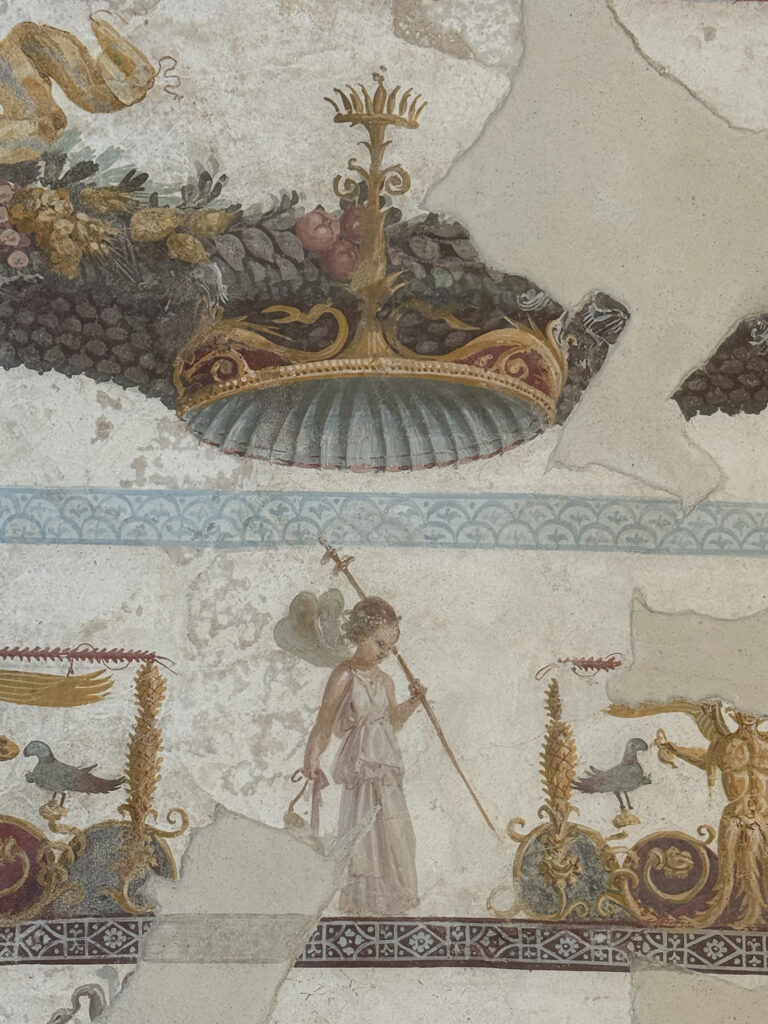A richly painted ceiling that once covered an upstairs banquet hall in Pompeii has been partially reconstructed from hundreds of fragments recovered beneath the room’s collapse. The restored section, from the House with the Bakery in Regio IX, reveals an elaborate mythological composition centred on the sacred union of Dionysus and Ariadne. Conservators have so far reassembled more than a quarter of the original vaulted ceiling, while additional pieces may still be identified during continuing excavations. The fragments were found where the upper floor of the house had fallen, allowing archaeologists to determine which room they originally decorated. Their positions were documented during excavation, and researchers began identifying connections between the pieces before conservation work formally started. Dionysus and Ariadne at the Centre The reconstructed decoration was arranged around a large central panel surrounded by architectural frames, floral garlands, winged figures, personifications of the seasons and other ornamental motifs. Although much of the main scene remains missing, comparisons with similar Roman artworks have helped specialists identify its subject. Dionysus is shown reclining on a chariot opposite Ariadne, who appears to be playing a harp. A winged Eros accompanies her with another stringed instrument, while two centaurs depicted as musicians pull the chariot. The scene represents the hierogamy, or sacred marriage, of Dionysus and Ariadne. References to the Dionysian world continue across the rest of the ceiling through theatrical masks, ritual vessels, drinking equipment and garlands filled with fruit and flowers. Such imagery would have been especially suited to a banquet room, where wine, music and performance formed part of Roman social life. Guests dining in the upstairs hall would have looked up at a composition celebrating Dionysus, the god associated with wine, theatre and ecstatic ritual. A Reconstruction Designed to Grow One of the most unusual aspects of the project is the support created for the surviving fragments. Instead of mounting the paintings on a conventional flat panel, conservators developed a curved structure that reproduces the shape of the original vault. Its exact form could only be established gradually as more fragments were joined together. The modular support also allows new pieces to be added without dismantling the existing reconstruction. This was necessary because other fragments linked to the ceiling remain in storage, while additional sections could emerge as archaeological work continues inside the house. Only pieces whose original positions could be established with sufficient confidence were included. Uncertain areas were left incomplete rather than being filled with hypothetical reconstructions. Pompeii Archaeological Park Director Gabriel Zuchtriegel said the project demonstrated how archaeological conservation could also become an opportunity for research and international collaboration. The ceiling adds to a series of recent discoveries connected with Dionysus in Regio IX. Between 2024 and 2025, excavations at the nearby House of the Thiasus uncovered a monumental painted procession associated with the god. Together, the two discoveries show how Dionysian mythology shaped some of Pompeii’s most richly decorated domestic interiors. In the House with the Bakery, however, the ancient image is still incomplete—and may continue to expand as more pieces of the collapsed ceiling are recovered and identified.