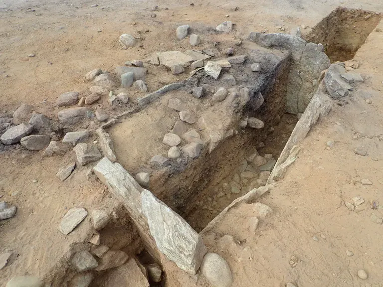 Overhead view of an excavated stone grave with large upright slabs along its sides and loose stones scattered around the opening.