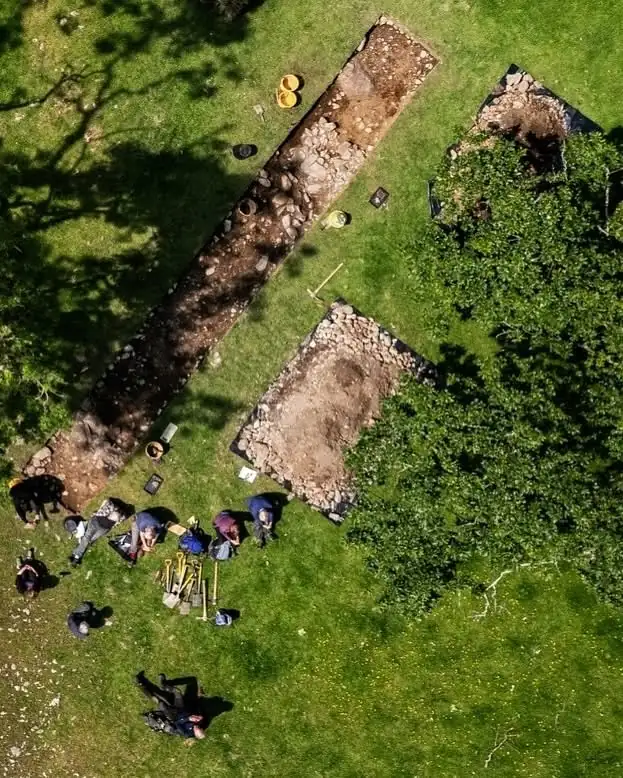 Aerial view of two excavation trenches in a grassy field, with volunteers working beside one trench.
