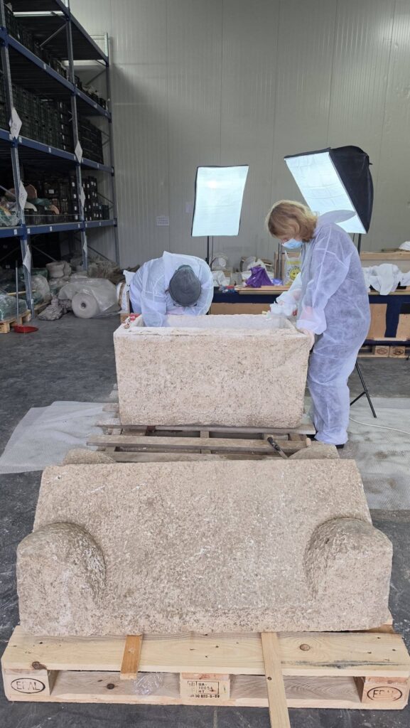 Archaeologists wearing protective clothing examining an opened Roman stone sarcophagus in a conservation facility.