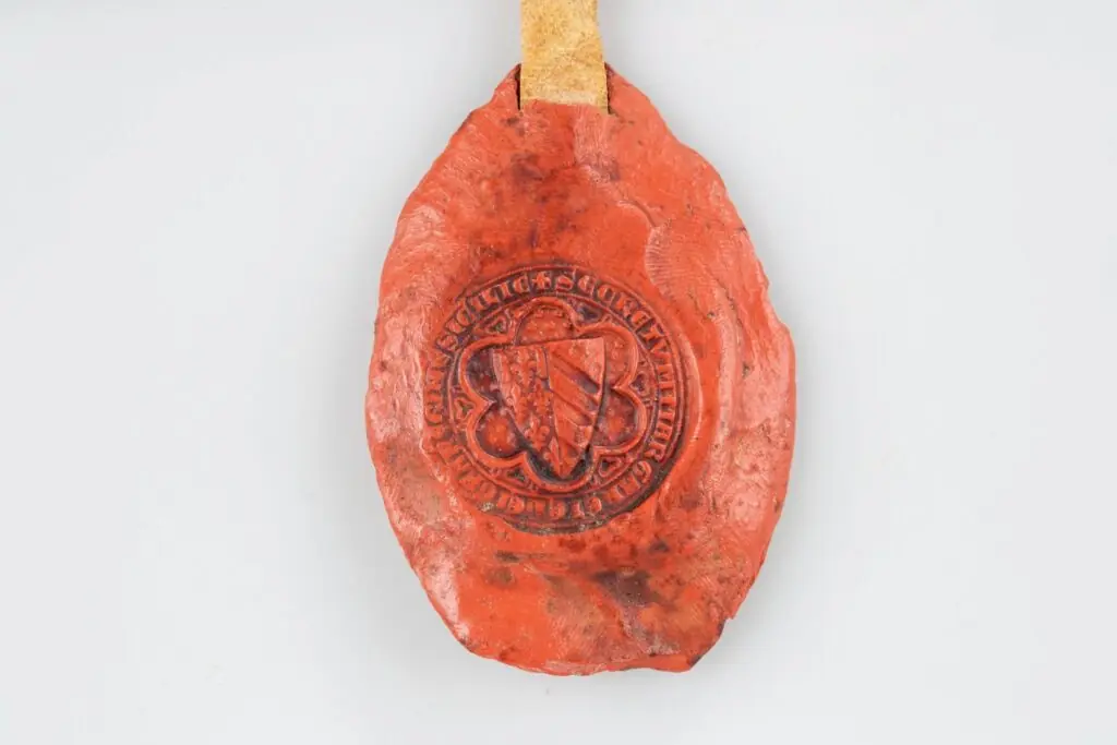 Detailed front view of a red medieval wax seal. A crowned queen stands in a Gothic architectural niche, holding a scepter, flanked by two small heraldic shields. A Latin inscription runs around the oval border.
