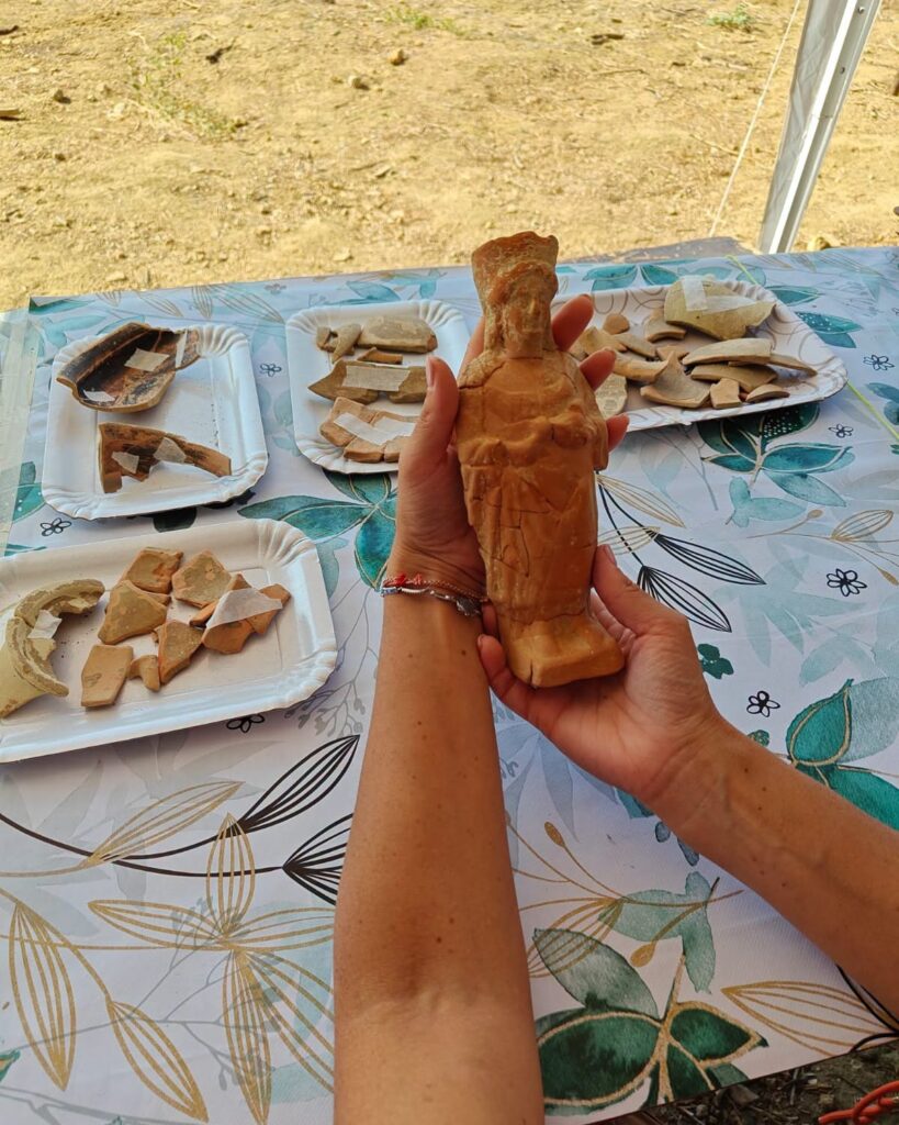 Ancient terracotta female figurine held in two hands beside pottery fragments at an archaeological excavation in Sicily.