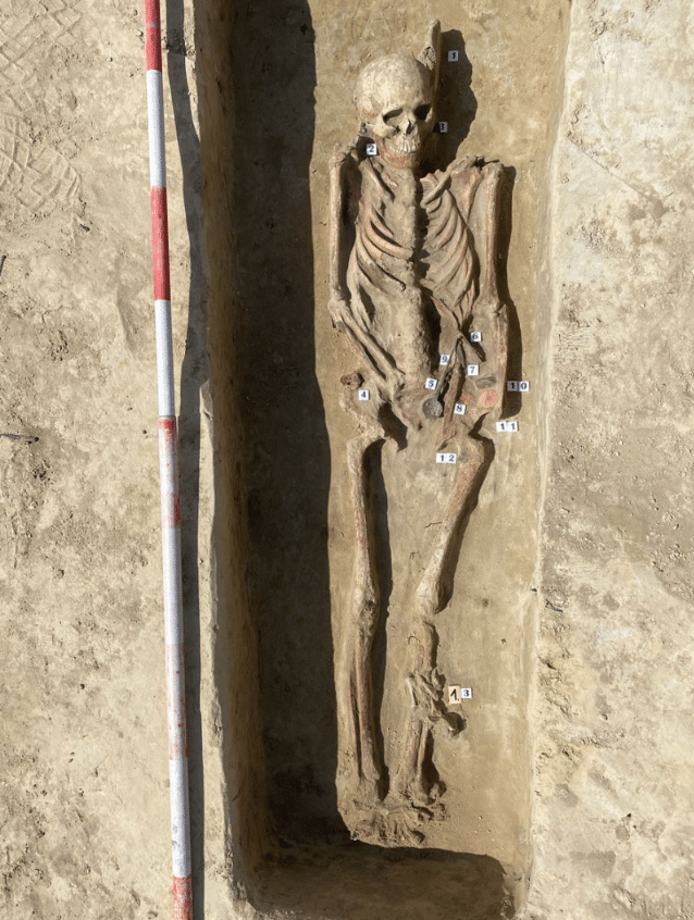 Excavated skeleton of an Avar woman surrounded by numbered archaeological finds in a narrow burial pit.