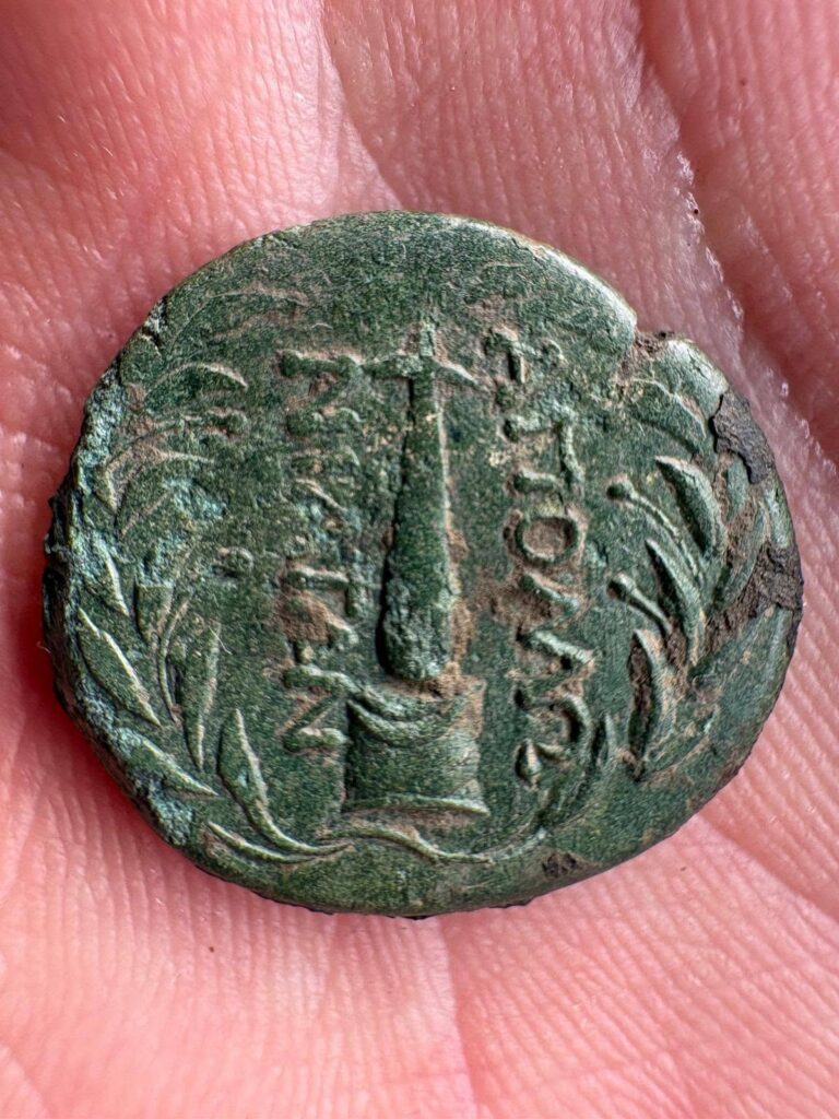 Close-up of an ancient bronze coin with green patina, Greek inscriptions, a central pillar-like motif and a surrounding wreath.