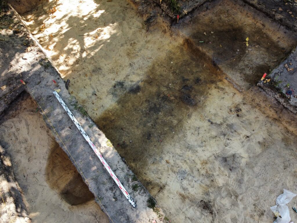 The excavation site of the early medieval boat-shaped cremation grave at Chodlik. The canoe-shaped grave, visible as a dark stain in the sand, and a meter stick next to it.