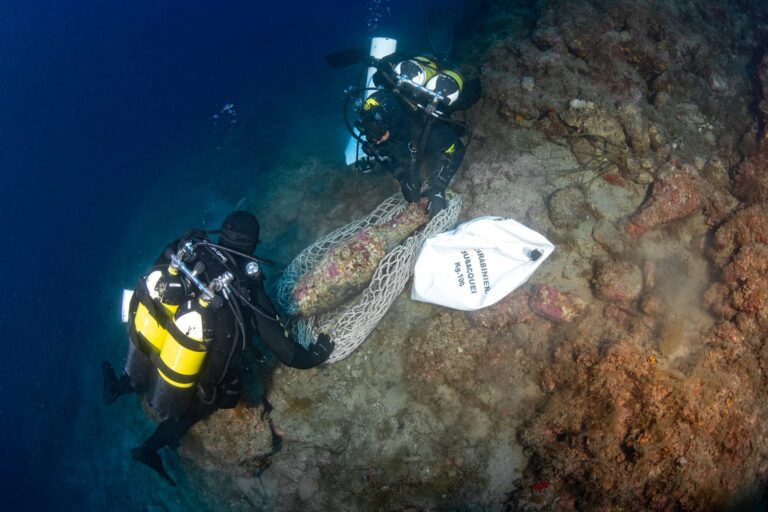 Two Carabinieri divers securing an ancient Roman amphora in a recovery net against an underwater rock face.