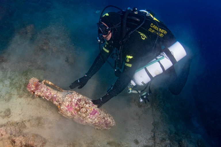 Underwater archaeologist examining a marine-encrusted Roman amphora among rocks near Punta Chiappa, Italy.