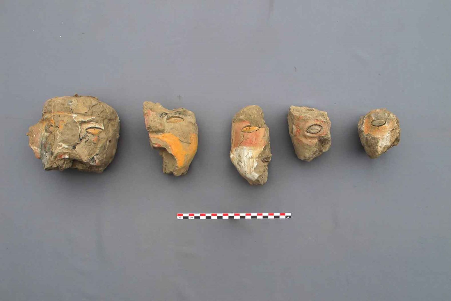 Five ancient Moche clay face fragments with surviving red, yellow, and black pigments arranged against a gray background.
