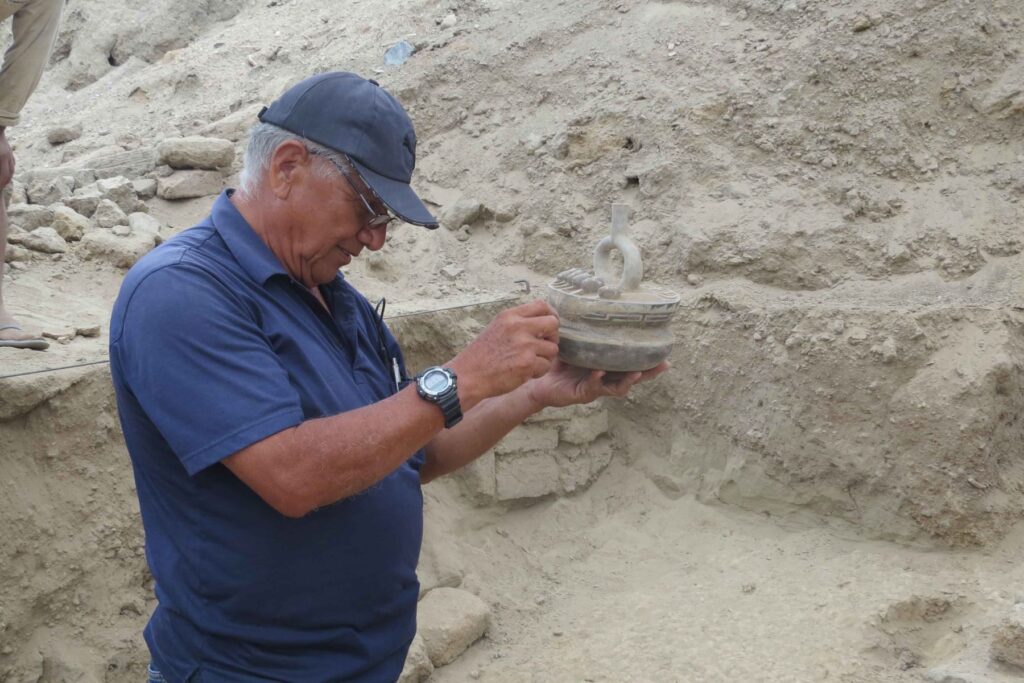 Man wearing a blue shirt and cap examining an ancient decorated ceramic vessel at an archaeological excavation site.