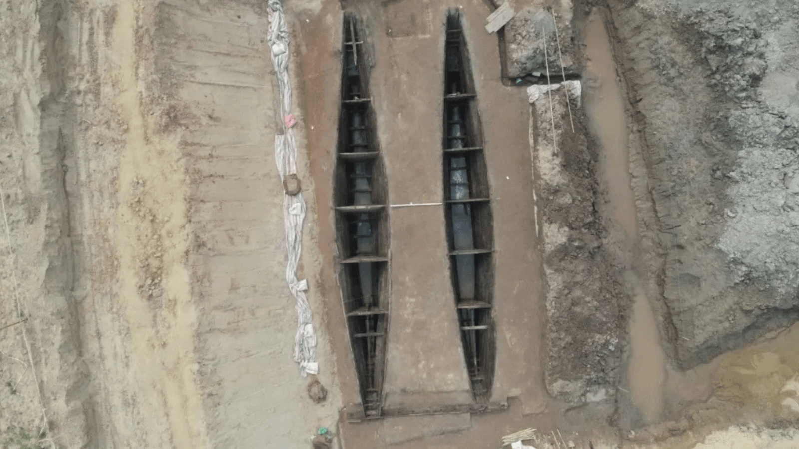 Overhead view of two parallel wooden hulls preserved in a muddy archaeological excavation.